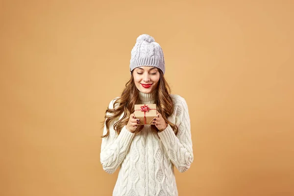 Ευτυχισμένος κορίτσι ντυμένοι με λευκό πλεκτό πουλόβερ και καπέλο κρατά ένα χριστουγεννιάτικο δώρο σε μπεζ φόντο στο στούντιο — Φωτογραφία Αρχείου