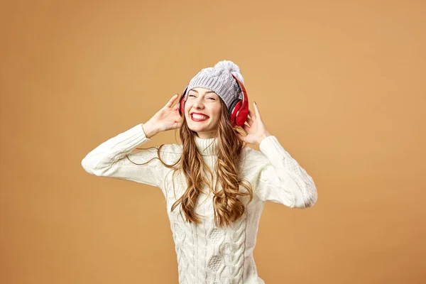 Ευτυχής κορίτσι κόκκινα ακουστικά ντυμένος με λευκό πλεκτό πουλόβερ και καπέλο διασκεδάζοντας σε μπεζ φόντο στο στούντιο — Φωτογραφία Αρχείου