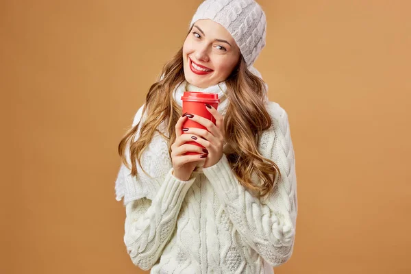 Усміхнена дівчина одягнена в білий в'язаний светр і капелюх тримає червону чашку в руках на бежевому фоні в студії — стокове фото