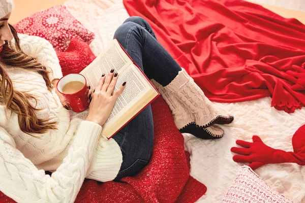 Une jeune fille vêtue d'un pull tricoté blanc, d'un jean et de chaussettes tricotées lit un livre couché sur des couvertures et des oreillers rouge-blanc avec une tasse de boisson chaude rouge et une assiette de biscuits — Photo