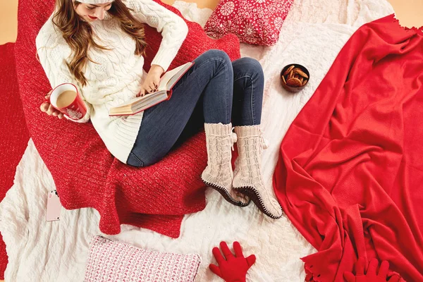 Дівчина в білому в'язаному светрі, джинсах і в'язаних шкарпетках лежить на червоно-білих ковдрах і подушках з червоною чашкою гарячого напою і тарілкою печива — стокове фото