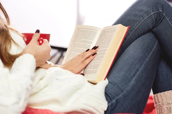 Девушка, одетая в белый вязаный свитер, джинсы и вязаные носки читает книгу и держит красную чашку горячего напитка в ней было — стоковое фото