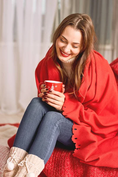 Дівчина, одягнена в білий в'язаний светр і джинси, вкриті червоною обгорткою, сидить з червоною чашкою гарячого напою в руках — стокове фото