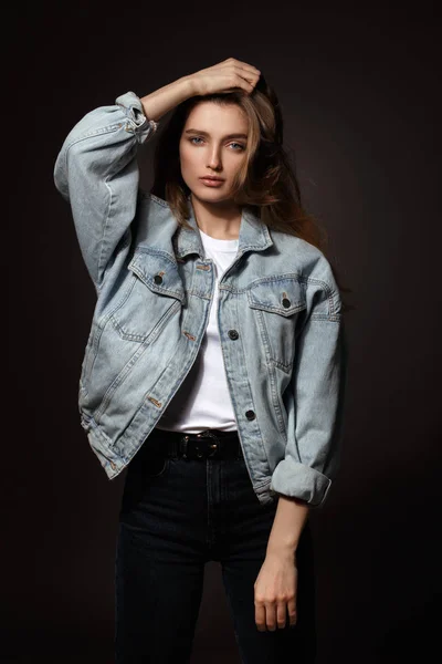 Gorgeous brunette girl with long flowing hair dressed in jeans jacket and jeans poses standing on the dark background in the studio — Stock Photo, Image