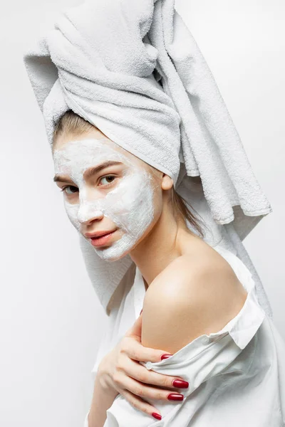 Beautiful young woman dressed in white clothes with a white towel on her hair and cosmetic mask on her face holds her hand on her naked shoulder — Stock Photo, Image
