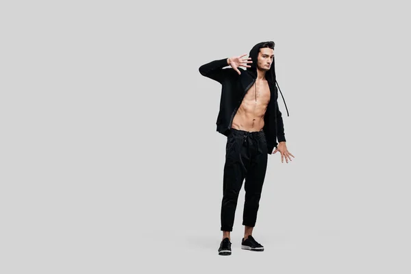 Dark-haired handsome dancer wearing sweatshirt on a naked torso and a black pants stands on a white background — Stock Photo, Image