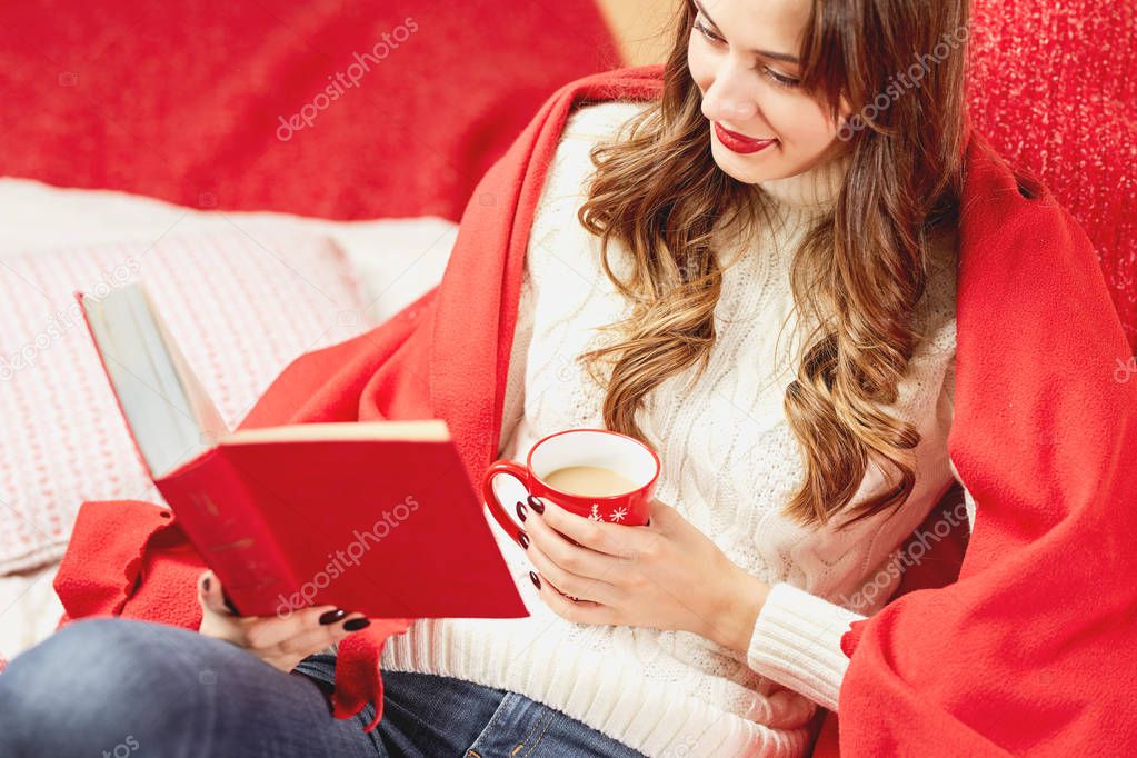 Girl dressed in white knitted sweater and jeans covered in red wrap is sitting with a red cup of hot drink and a book in her hands  on red-white blankets