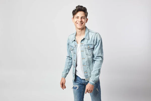 Sorrindo jovem de cabelos escuros cara em uma camiseta branca, jeans e uma jaqueta de ganga fica no fundo branco no estúdio — Fotografia de Stock