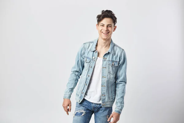 Sorrindo jovem de cabelos escuros cara em uma camiseta branca, jeans e uma jaqueta de ganga fica no fundo branco no estúdio — Fotografia de Stock