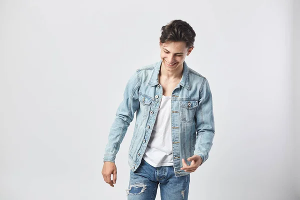 Sorrindo jovem de cabelos escuros cara em uma camiseta branca, jeans e uma jaqueta de ganga fica no fundo branco no estúdio — Fotografia de Stock