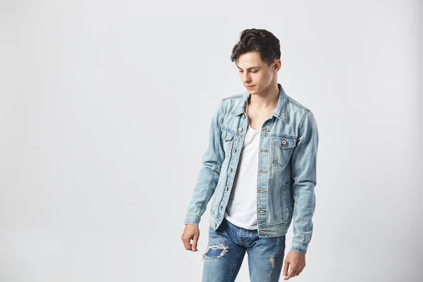 Jeune homme brun avec un t-shirt blanc, un jean et une veste en denim se tient sur le fond blanc dans le studio — Photo