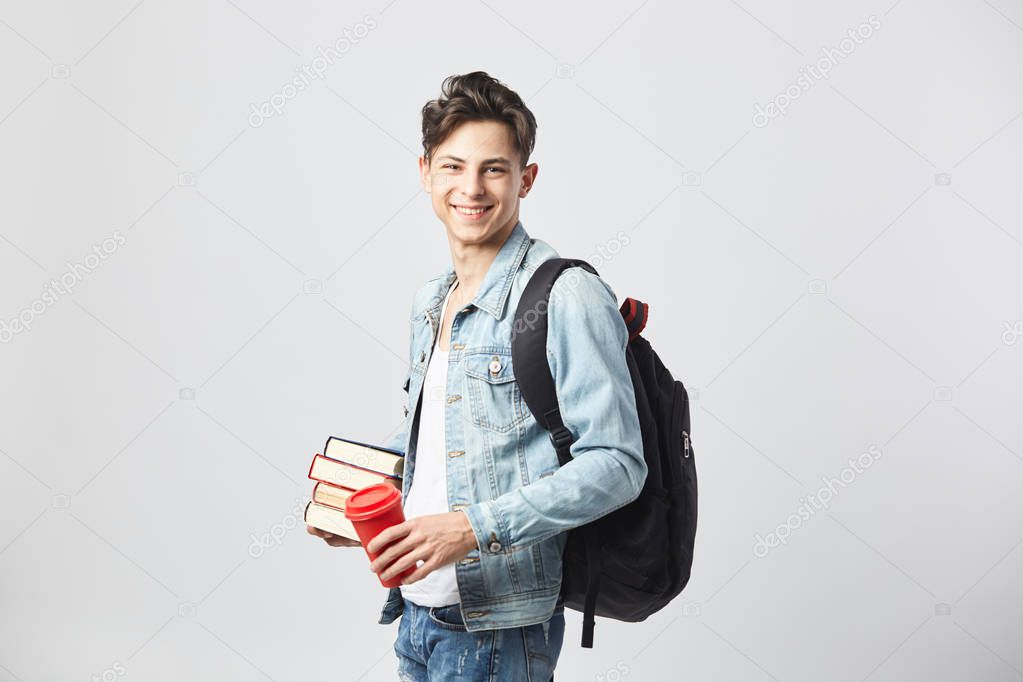 Smiling young dark-haired guy with black backpack on his shoulder dressed in a white t-shirt, jeans and a denim jacket holds books and red plastic cup in his hands on the white background in the