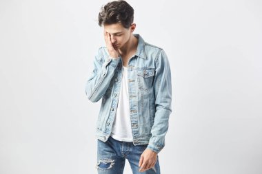 Beyaz t-shirt, kot pantolon ve bir kot ceket giymiş üzgün esmer adam stüdyo beyaz zemin üzerine yüzündeki elini tutar