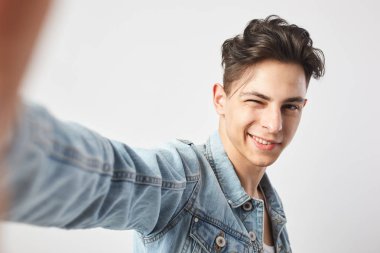 Esmer adam gülümseyen bir beyaz t-shirt giymiş ve bir kot ceket winks ve stüdyoda beyaz zemin üzerine bir salfie yapar