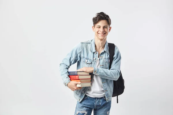 Sorrindo jovem de cabelos escuros cara com mochila preta em seu ombro vestido com uma camiseta branca, jeans e uma jaqueta de ganga contém livros em suas mãos sobre o fundo branco no estúdio — Fotografia de Stock