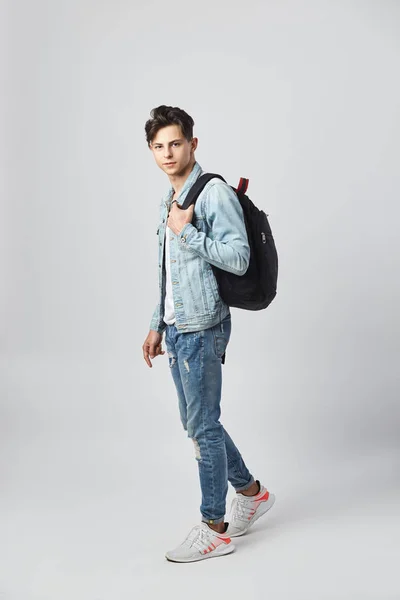 Mörkhårig kille med svart ryggsäck på hans axel klädd i en vit t-shirt, jeans och en jeansjacka står på den vita bakgrunden i studion — Stockfoto