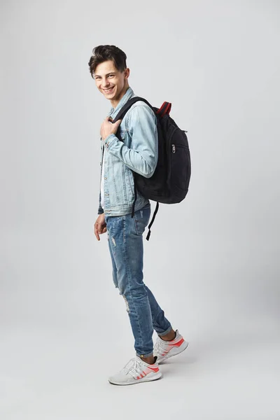 Leende ung mörkhårig kille med svart ryggsäck på hans axel klädd i en vit t-shirt, står jeans och en jeansjacka på den vita bakgrunden i studion — Stockfoto