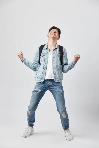 Jonge dark-haired kerel met zwarte rugzak op zijn schouder gekleed in een wit t-shirt, jeans en een denim jasje ziet er gelukkig op de witte achtergrond in de studio — Stockfoto