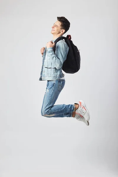 Jovem de cabelos escuros com mochila preta no ombro vestida com uma camiseta branca, jeans e uma jaqueta de ganga salta alegremente no fundo branco do estúdio — Fotografia de Stock