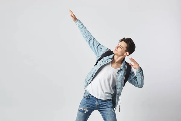 Ung mörkhårig kille med svart ryggsäck på hans axel klädd i en vit t-shirt, jeans och en jeansjacka höjde ena handen upp mot den vita bakgrunden i studion — Stockfoto