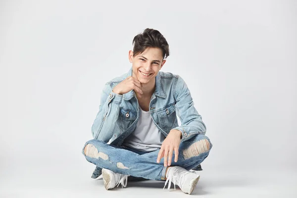 Un homme souriant aux cheveux bruns vêtu d'un t-shirt blanc, d'un jean et d'une veste en jean est assis les jambes croisées sur le sol sur le fond blanc du studio — Photo