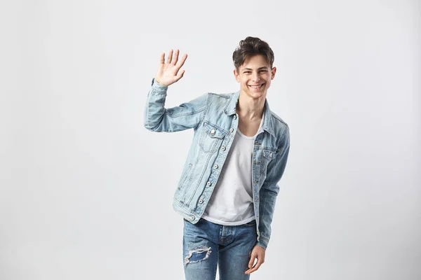 Amigável cara de cabelos escuros vestido com uma camiseta branca, jeans e uma jaqueta de ganga fica no fundo branco no estúdio — Fotografia de Stock