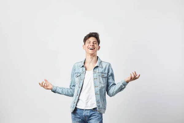 Rindo cara de cabelos escuros vestido com uma camiseta branca, jeans e uma jaqueta de ganga abre os braços para o lado no fundo branco no estúdio — Fotografia de Stock