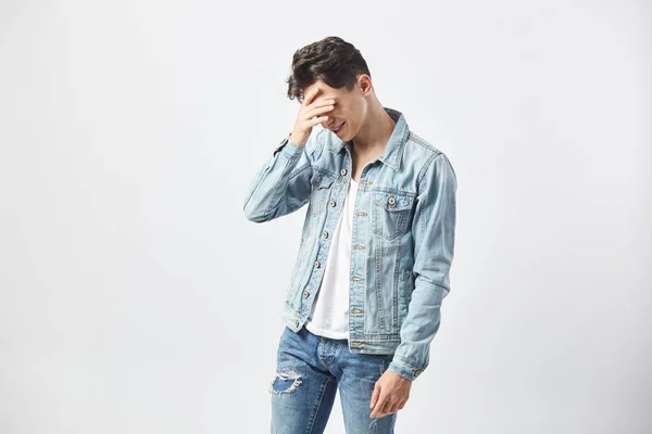 Smutné, černovlasý muž, oblečený v bílé tričko, džíny a džínové bundy drží ruku na tvář na bílém pozadí v ateliéru — Stock fotografie