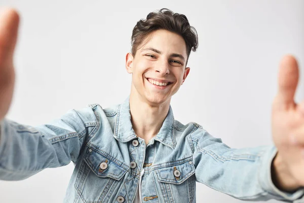 Sorrindo cara de cabelos escuros vestido com uma camiseta branca e uma jaqueta de ganga faz uma salfie no fundo branco no estúdio — Fotografia de Stock