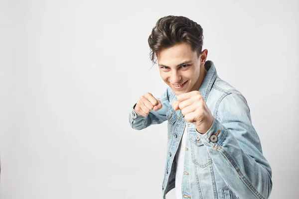 Ung mörkhårig kille klädd i en vit t-shirt och en jeansjacka håller händerna i nävarna på den vita bakgrunden i studion — Stockfoto