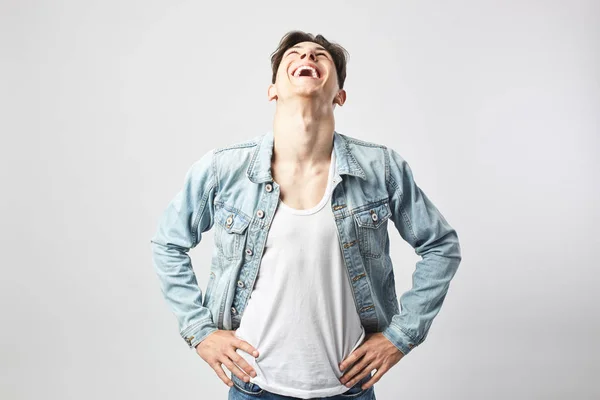 Ragazzo ridente ragazzo dai capelli scuri vestito con una t-shirt bianca e una giacca di jeans si erge sullo sfondo bianco in studio — Foto Stock