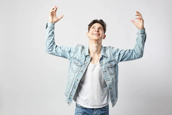 Smiling guy dark-haired guy dressed in a white t-shirt and a denim jacket stands and raises his hands up on the white background in the studio