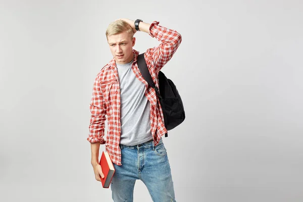Blond kille med svart ryggsäck på hans axel klädd i en vit t-shirt, röd rutig skjorta och jeans håller boken i sin hand och andra handen på huvudet på den vita bakgrunden i studion — Stockfoto