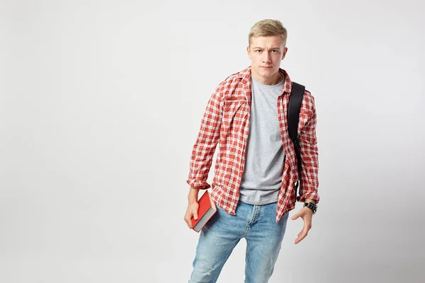 Allvarlig blond kille med svart ryggsäck på hans axel klädd i en vit t-shirt, röd rutig skjorta och jeans håller boken i sin hand på den vita bakgrunden i studion — Stockfoto