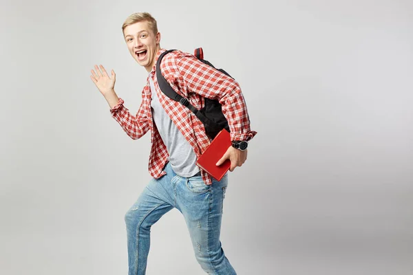 Joyeux blond avec un sac à dos noir sur l'épaule habillé d'un t-shirt blanc, chemise à carreaux rouges et un jean tient le livre dans sa main et crie sur le fond blanc dans le studio — Photo