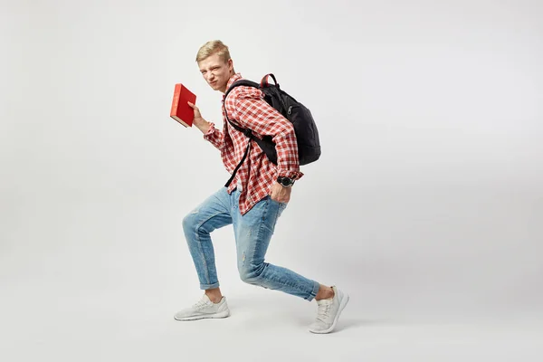 Glada blond kille med svart ryggsäck på hans axel klädd i en vit t-shirt, röd rutig skjorta och jeans håller boken i sin hand och ropar på den vita bakgrunden i studion — Stockfoto