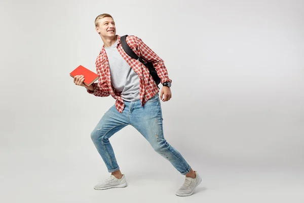 Joyful blond guy with black backpack on his shoulder dressed in a white t-shirt, red checkered shirt and jeans holds book in his hand and shouts on the white background in the studio