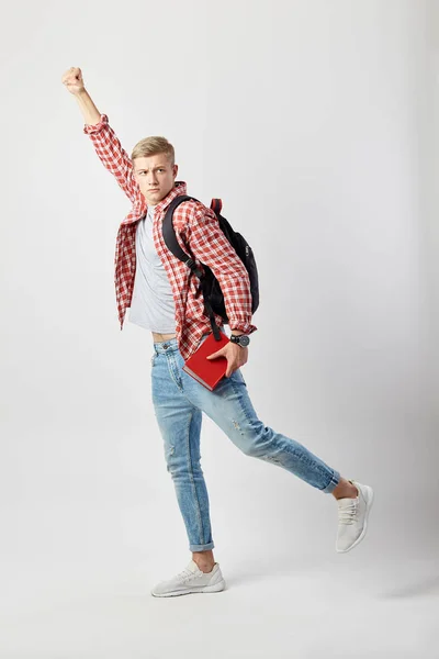 Rolig blond kille med svart ryggsäck på hans axel klädd i en vit t-shirt, röd rutig skjorta och jeans håller boken i sin hand att höja sin andra hand på den vita bakgrunden i studion — Stockfoto