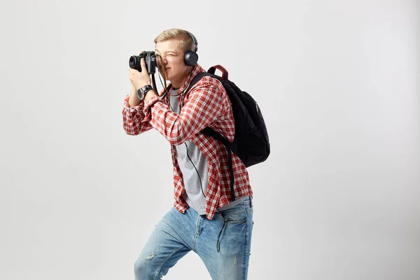 Blond kille i hörlurar, med svart ryggsäck på hans axel klädd i en vit t-shirt, röd rutig skjorta och jeans gör bilder på kameran på den vita bakgrunden i studion — Stockfoto