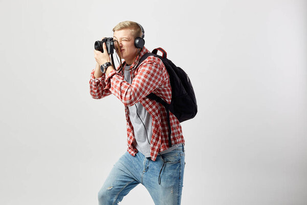 Blond guy in headphones, with black backpack on his shoulder dressed in a white t-shirt, red checkered shirt and jeans makes photos on camera on the white background in the studio