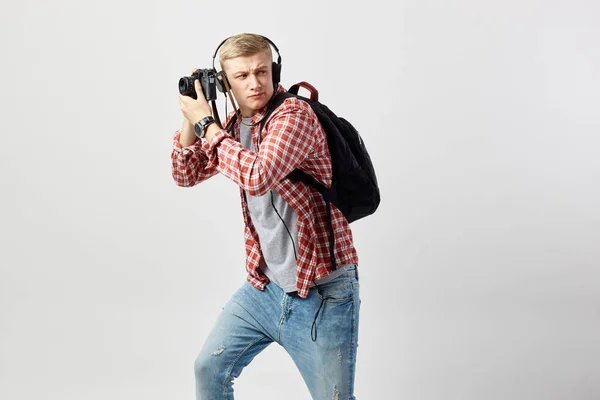 Blond kille i hörlurar, med svart ryggsäck på hans axel klädd i en vit t-shirt, röd rutig skjorta och jeans gör bilder på kameran på den vita bakgrunden i studion — Stockfoto