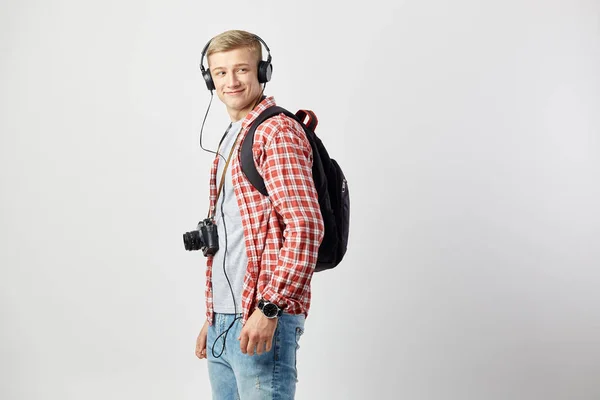 Blond kille i hörlurar, med svart ryggsäck på hans axlar klädd i en vit t-shirt, röd rutig skjorta och jeans står på den vita bakgrunden i studion med kameran på halsen — Stockfoto