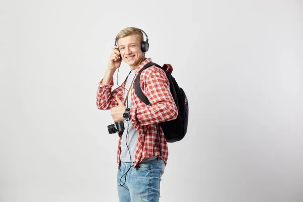 Blond kille i hörlurar, med svart ryggsäck på hans axlar klädd i en vit t-shirt, röd rutig skjorta och jeans står på den vita bakgrunden i studion med kameran på halsen — Stockfoto