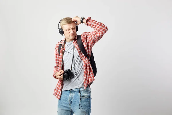 Blond kille i hörlurar, med svart ryggsäck på hans axel klädd i en vit t-shirt, röd rutig skjorta och jeans håller kameran i händerna på den vita bakgrunden — Stockfoto