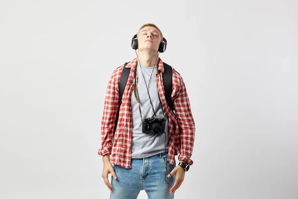 Blond kille i hörlurar, med svart ryggsäck på hans axlar klädd i en vit t-shirt, röd rutig skjorta och jeans står på den vita bakgrunden i studion med kameran på halsen — Stockfoto