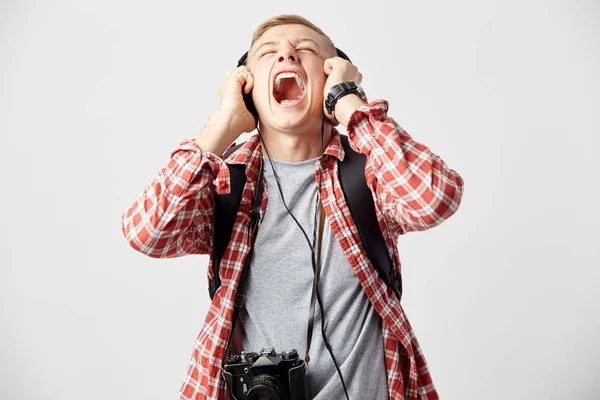 Blond kille i hörlurar, med svart ryggsäck på hans axlar klädd i en vit t-shirt och röd rutig skjorta ropar i studion med kameran på halsen — Stockfoto