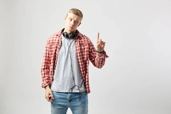 Blond kille med hörlurar på halsen klädd i en vit t-shirt, röd rutig skjorta och jeans står på den vita bakgrunden i studion och punkt upp med fingret — Stockfoto