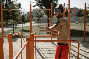 Atletik çıplak gövde ile kafa bandı erkekte siyah tayt giymiş ve kırmızı şort dışında spor zeminde asimetrik paralel yanındaki güneşli bir günde germe yapıyor