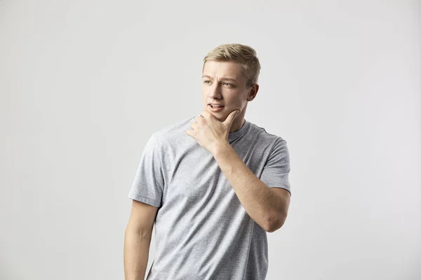 Blondýna chlap oblečený v bílé tričko stojí na bílém pozadí v ateliéru, držela ho za ruku na bradě — Stock fotografie