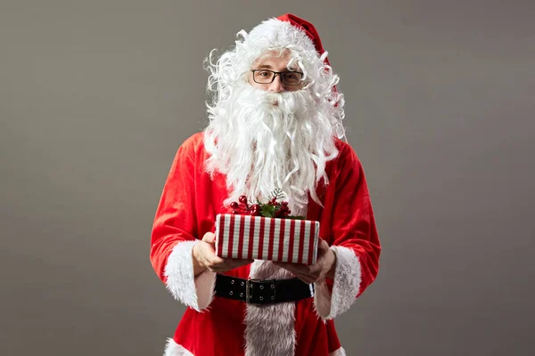 Санта-Клаус в окулярах тримає в руці Різдвяний подарунок на сірий фон. — стокове фото